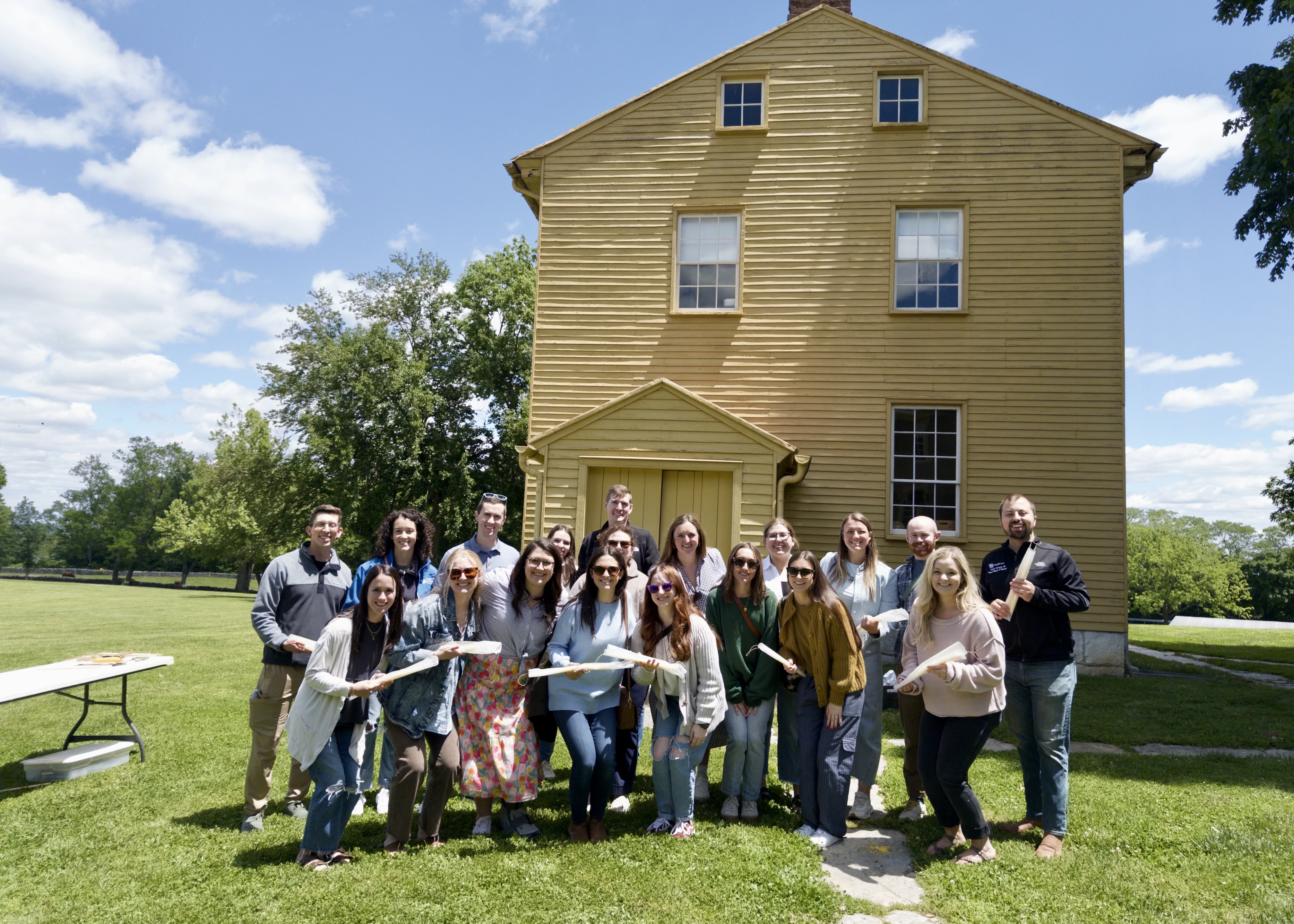 A cheerful group poses on a sunny lawn in front of a large historic yellow wooden house, holding a long white banner amid green trees and blue sky.