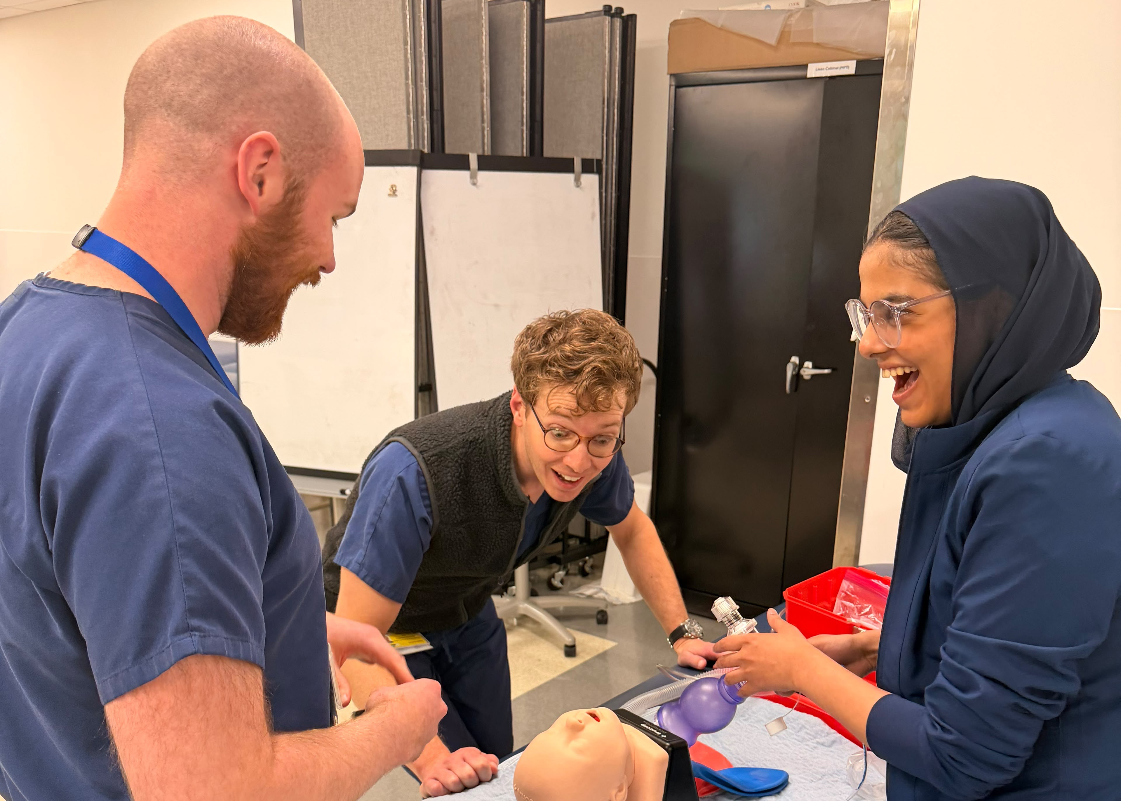Three healthcare trainees practice on a training dummy in a classroom, smiling as they work together with a purple resuscitation bag, creating a warm, collaborative scene.