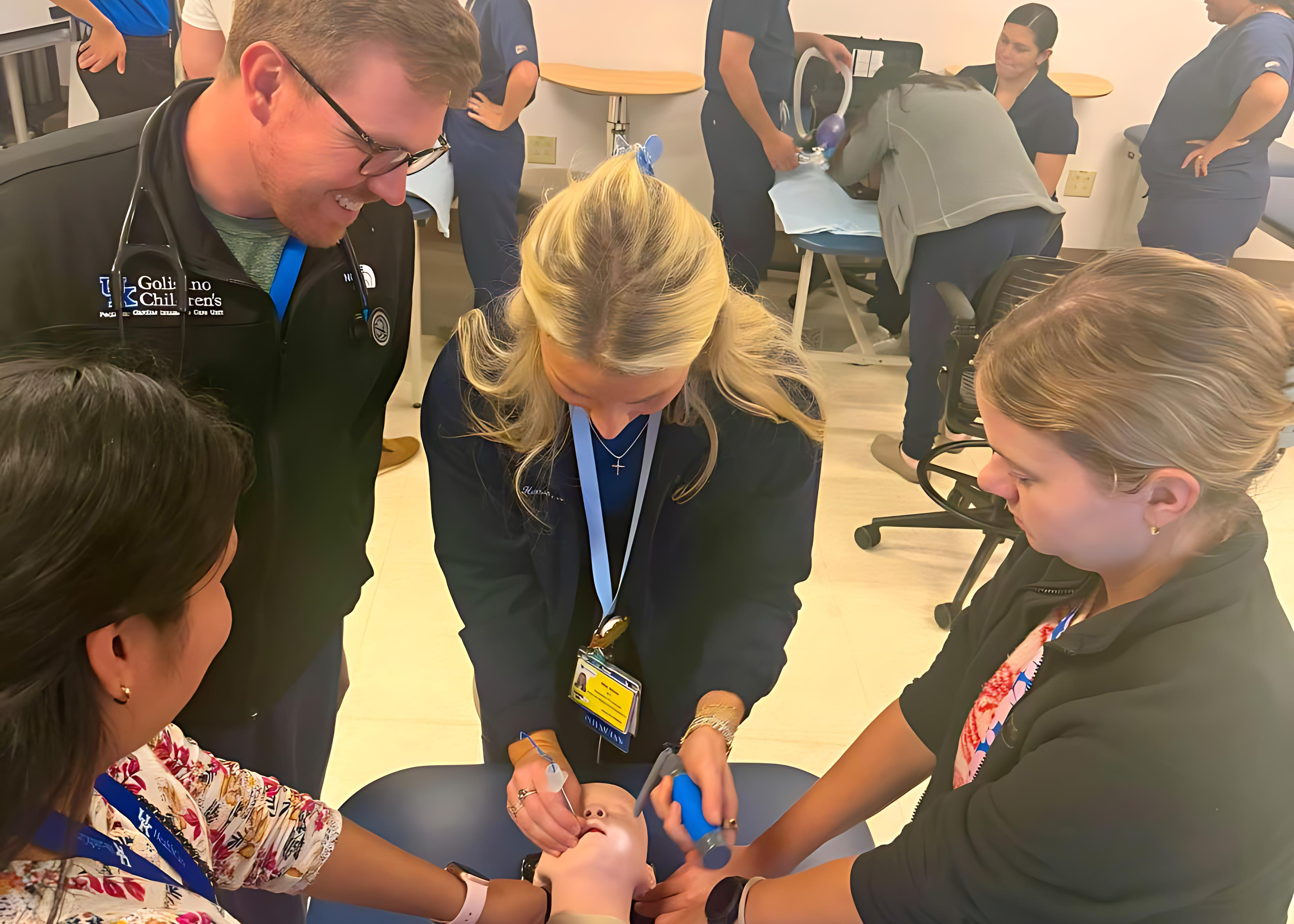 Medical staff gather around a simulation dummy as a clinician demonstrates a blue medical device on their hand, creating a focused, supportive training scene.