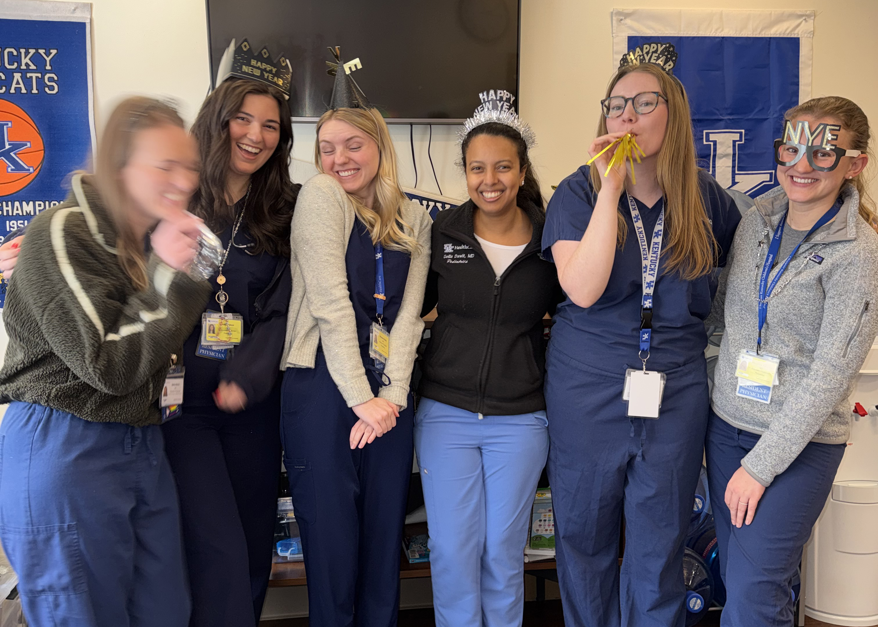 Six women in a team room wear paper crowns and lanyards; one blows a party horn as friends smile, with sports banners behind them.