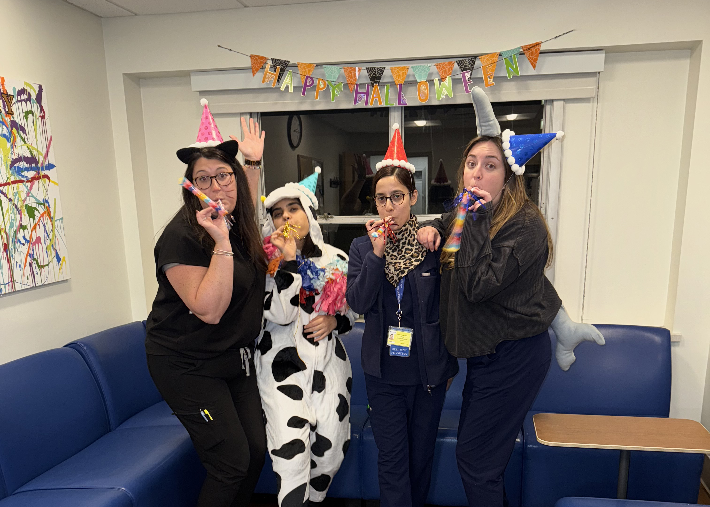 Four women in playful Halloween costumes pose with party blowers beneath a “Happy Halloween” banner in an office lounge, smiling in a festive mood.