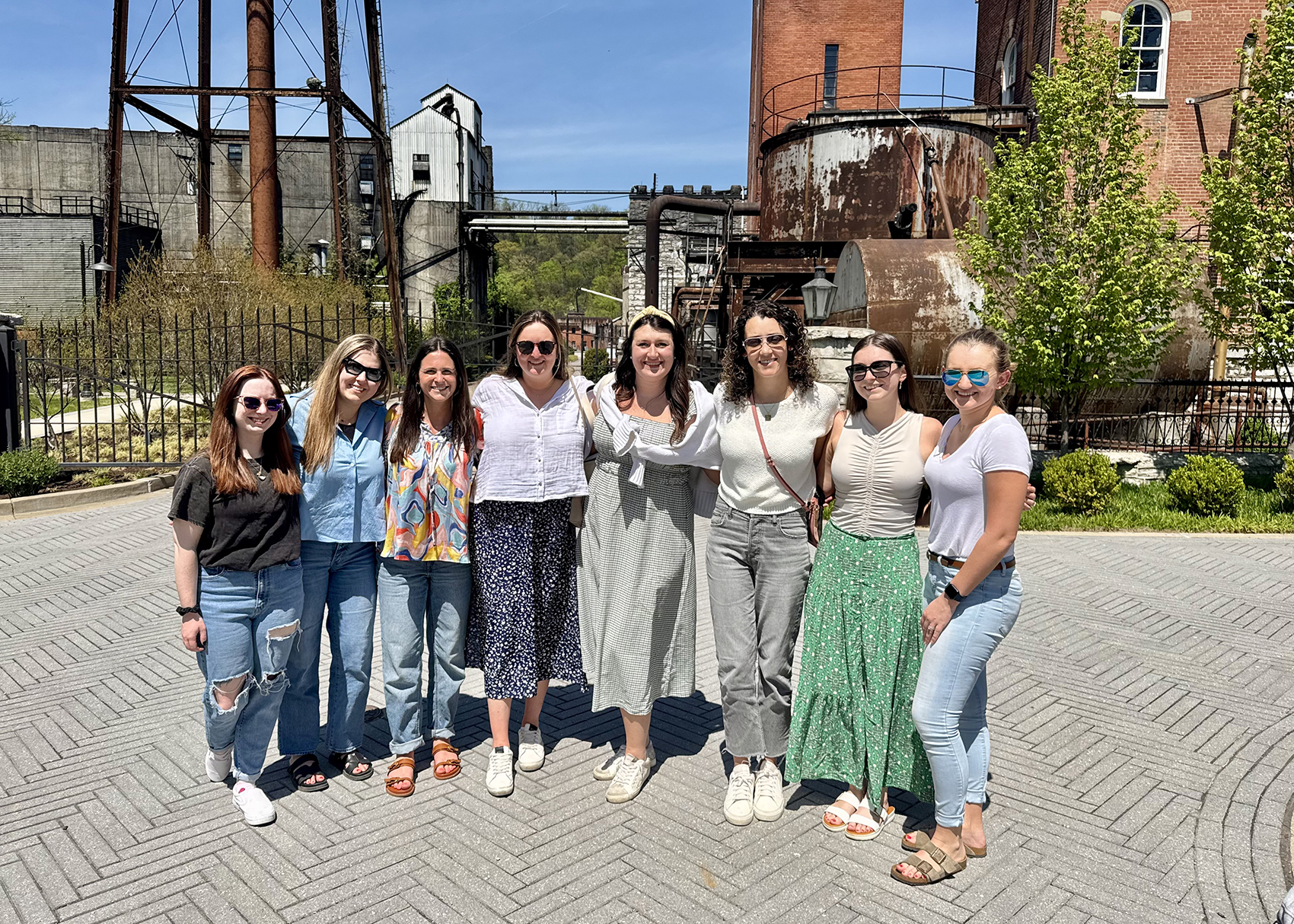 Eight women pose together on a sunny paved walkway at a historic industrial site, with brick smokestacks and metal structures behind them; the mood is cheerful and relaxed.