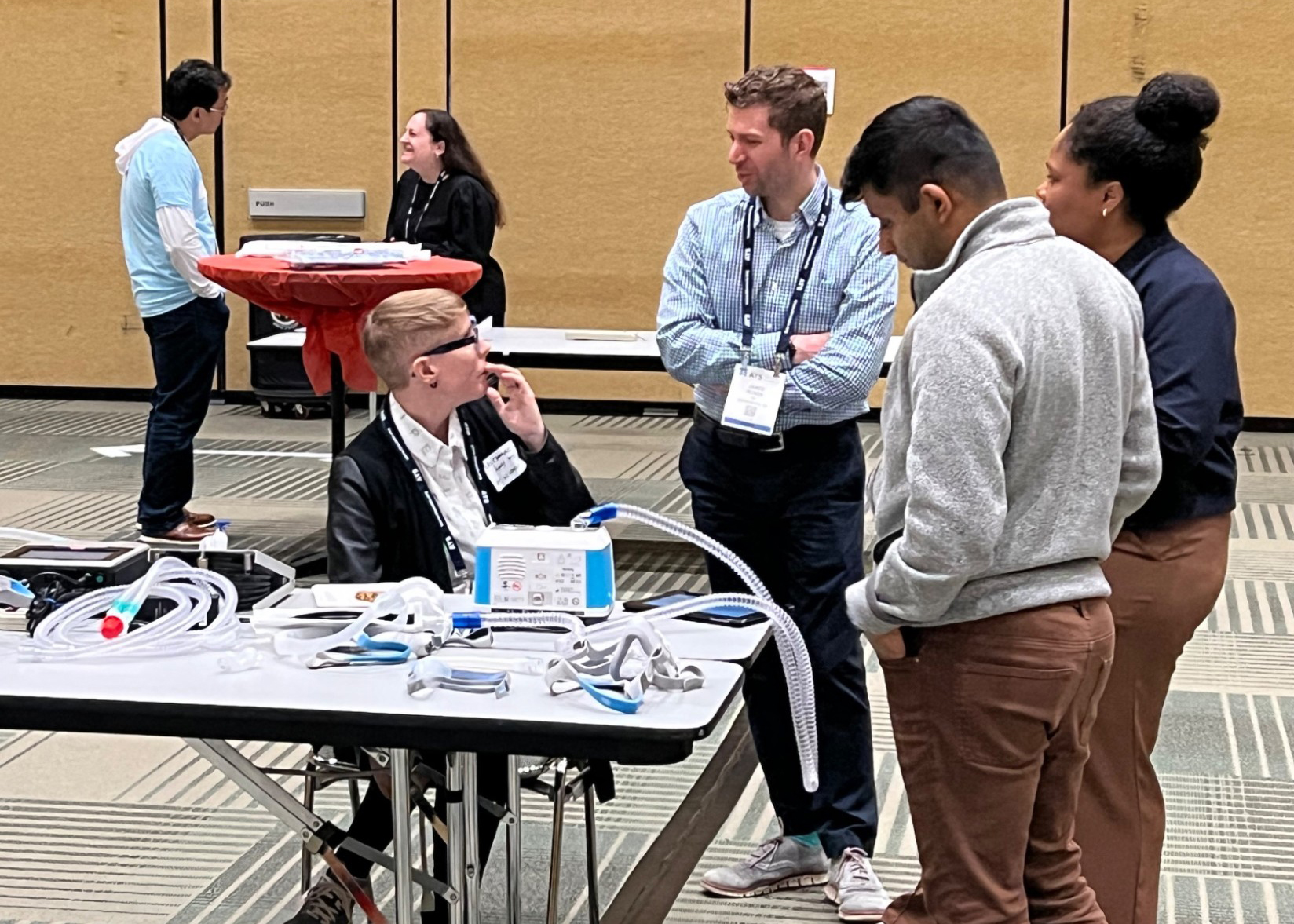 At a medical equipment demonstration, a seated presenter explains devices and tubing on a table to four attentive attendees in a conference room.