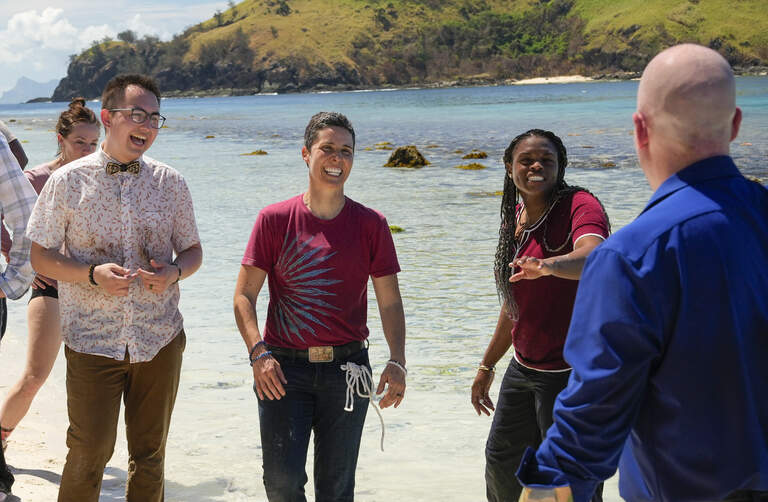 Still from Survivor episode, A cheerful group chats in shallow tropical water beside a green island, with a bald man in blue facing three smiling companions; relaxed, friendly mood.