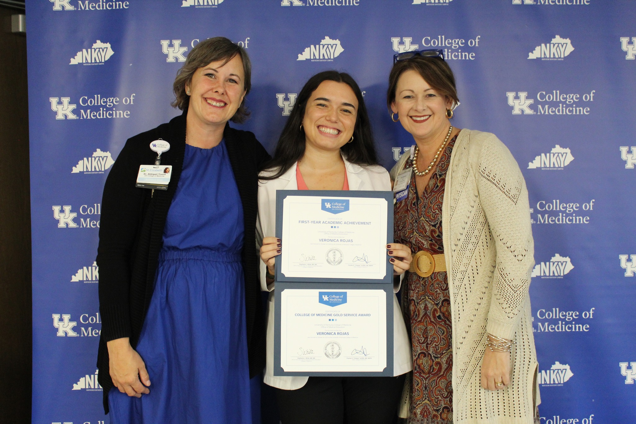 Awardee and presenters hold two certificates and pose for a photo in front of blue backgroudn