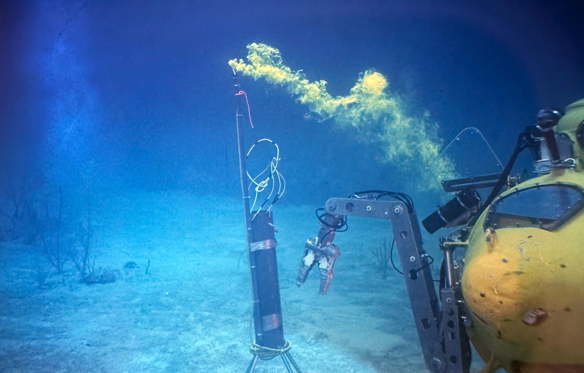 Underwater scene showing a yellow submersible's robotic arm interacting with a submerged object releasing yellowish plumes, set against a blue ocean floor. | Photo provided by Dr. Gerhardt