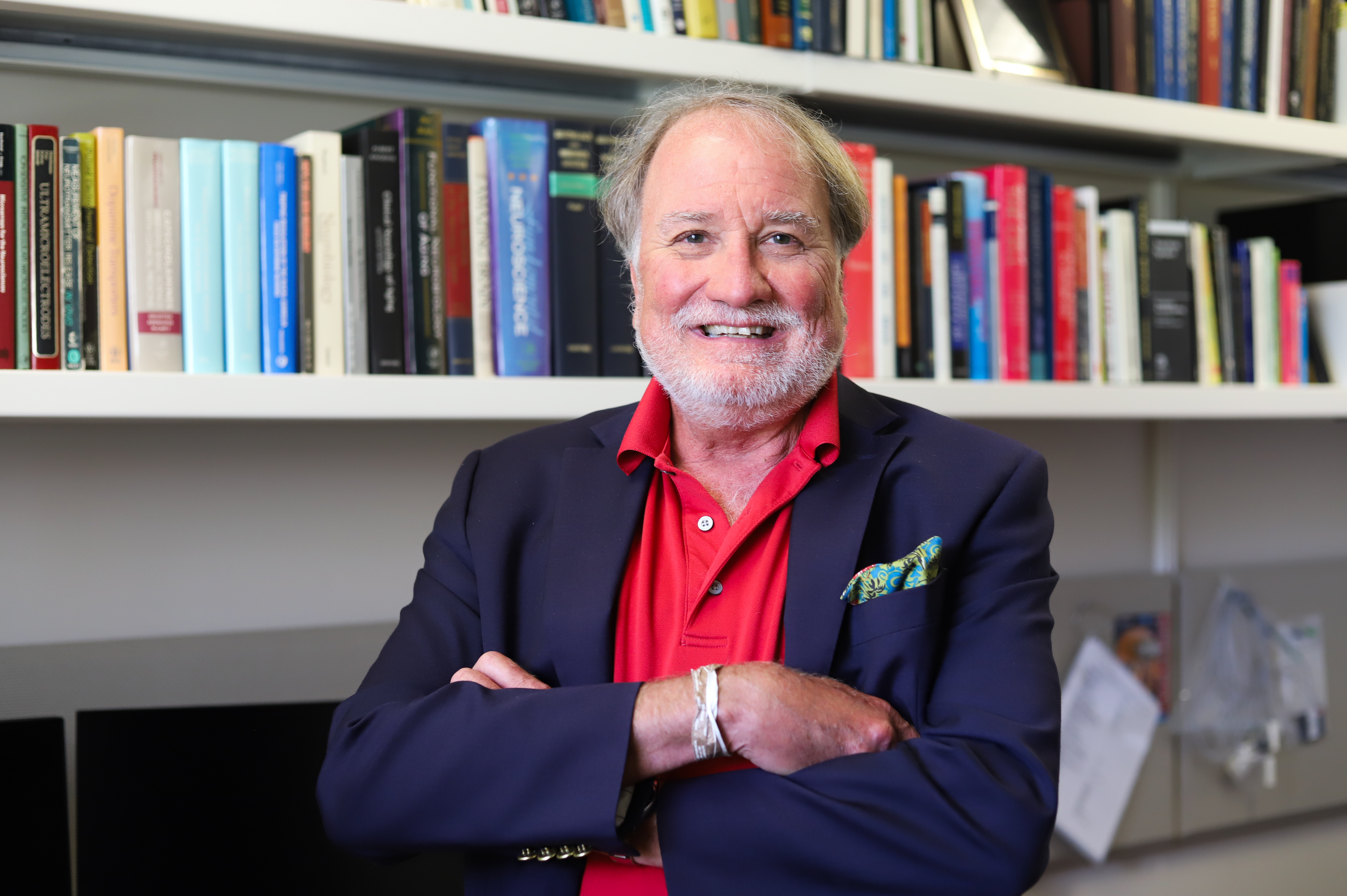 Dr. Gerhardt with crossed arms, wearing a navy blazer and red shirt, stands in front of a bookshelf filled with colorful books in an office setting. | Photo by Lily Wilder