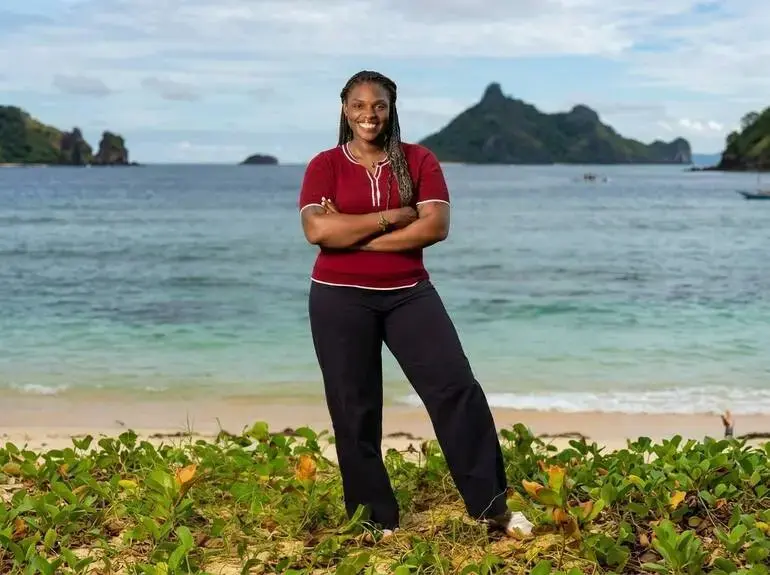 Promotional photo of Sharonda Cox from Survivor, A smiling woman in a red shirt and black trousers stands with arms crossed on a tropical beach, with turquoise water, green islands, and a bright sky behind her.
