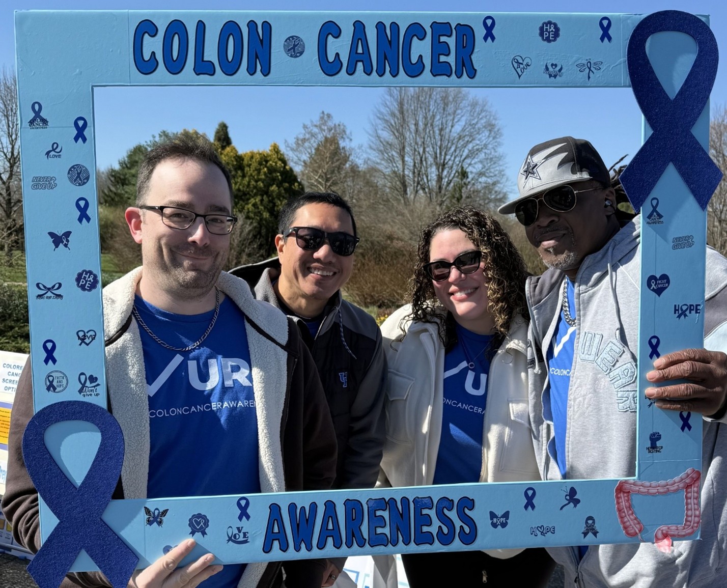 In a large cardboard frame that reads "Colon Cancer Awareness" four people smile, holding up the frame.