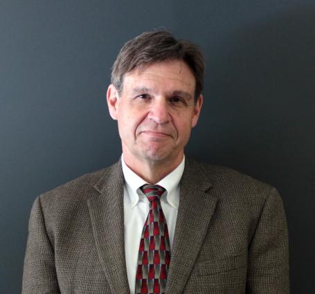 Dr. Tom Kelly wears a brown suit and maroon tie, in front of a gray wall