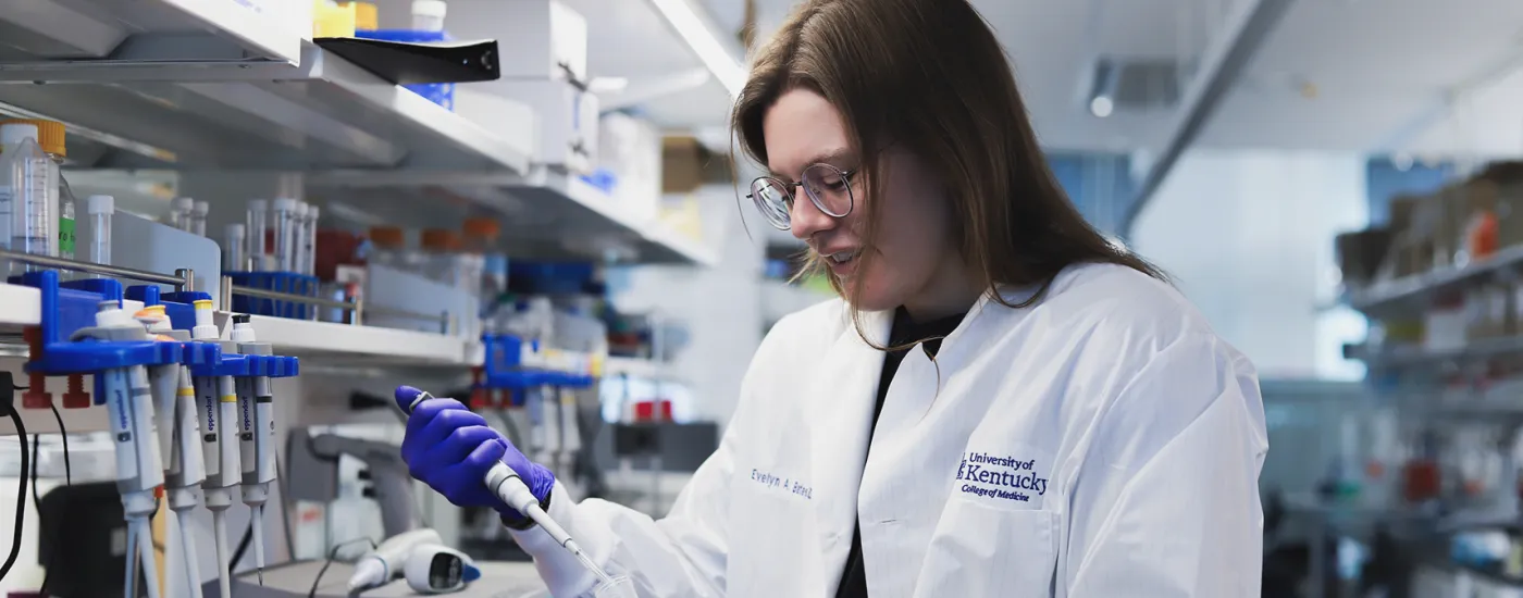 A University of Kentucky College of Medicine research trainee and PhD student works in a laboratory, wearing a white lab coat and purple gloves while using a pipette. She is surrounded by lab equipment, including pipettes and sample tubes, indicating an active research environment.