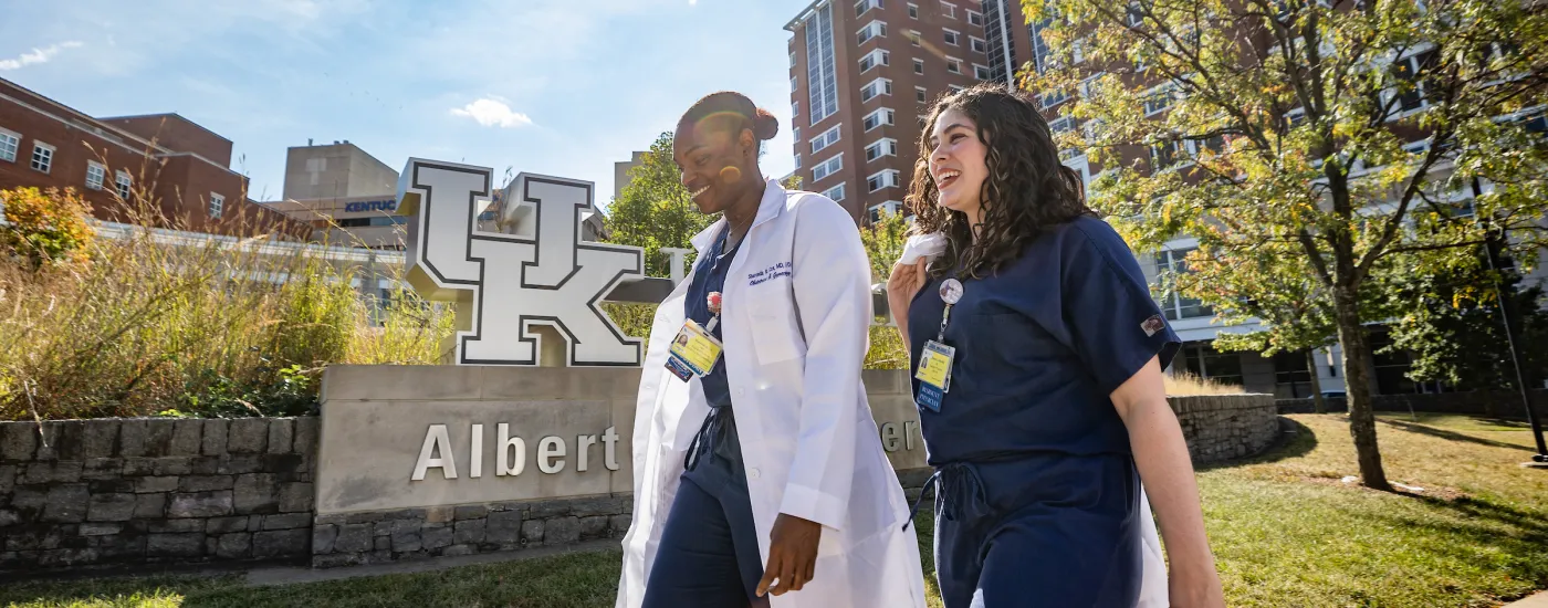 two residents, one in scrubs and one in a white coat, walk outside in front of the uk chandler hospital with the UK logo visible in the background