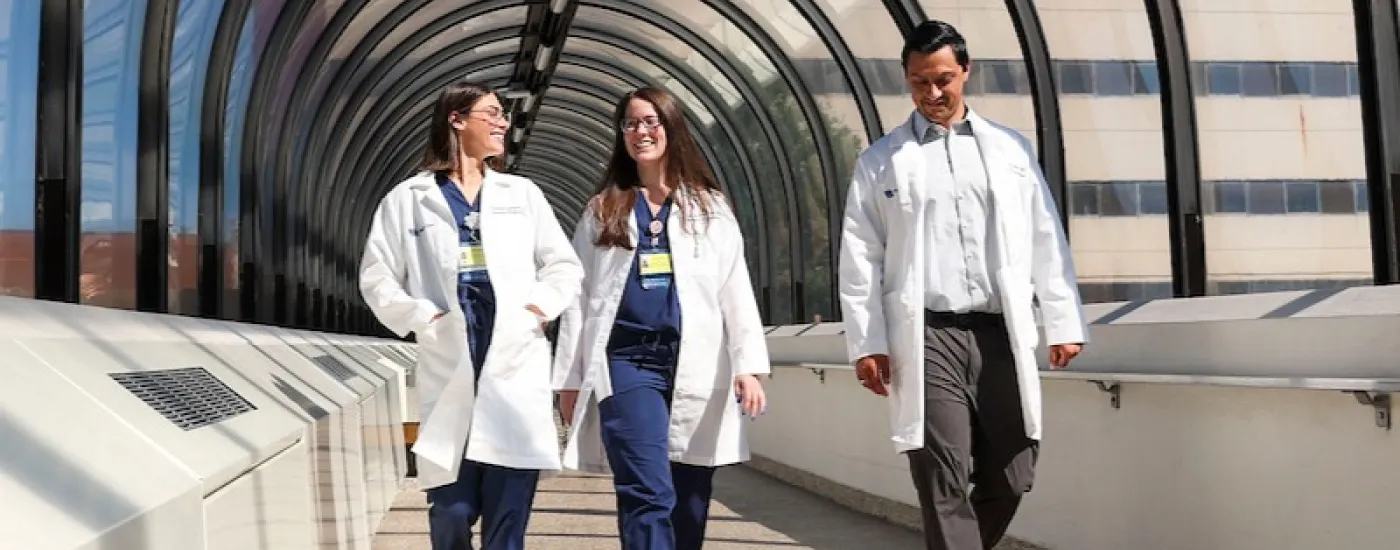 Two women in white coats and blue scrubs and one man in a white coat, white shirt, and brown pants walking side by side in a well-lit pedestrian tunnel
