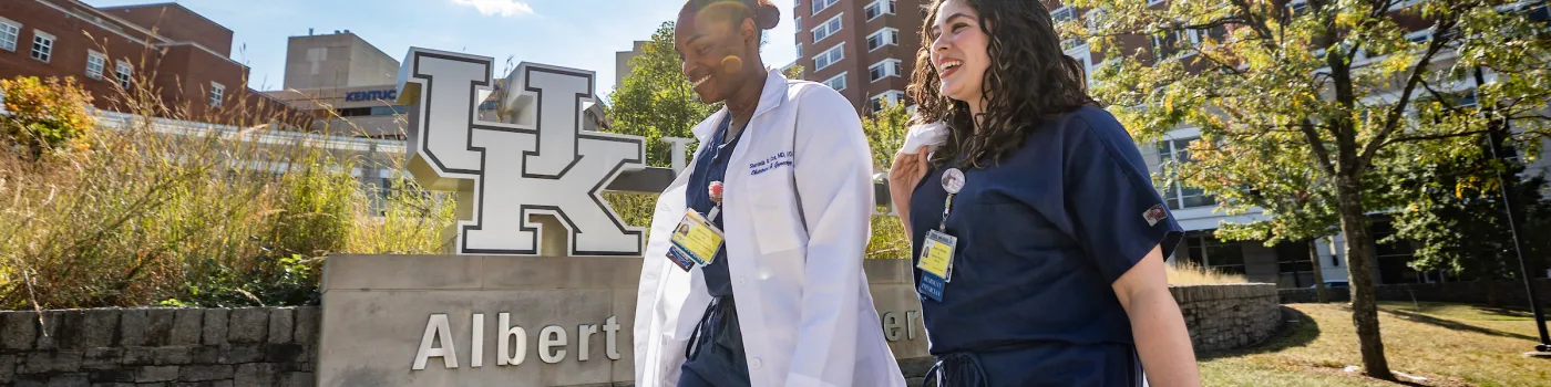 two residents, one in scrubs and one in a white coat, walk outside in front of the uk chandler hospital with the UK logo visible in the background