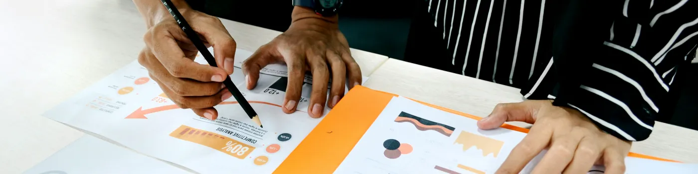 Two people hover a pen over various sheets of notes and graphs