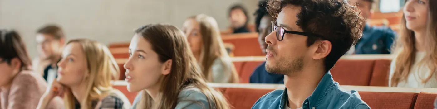 Students attentively listen in a lecture hall. They sit in rows, taking notes. The atmosphere is focused and engaged, suggesting a learning environment.