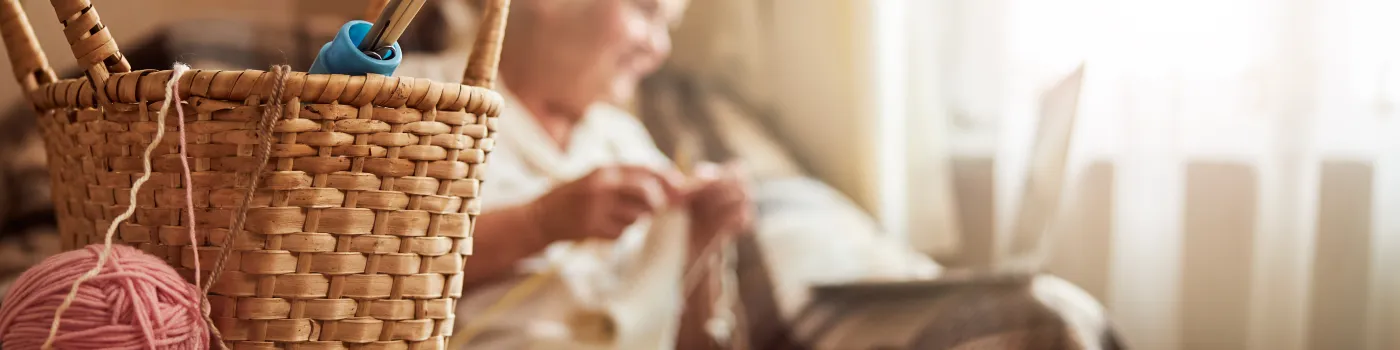 An elderly woman is knitting with a basket full of yarn on the table. 