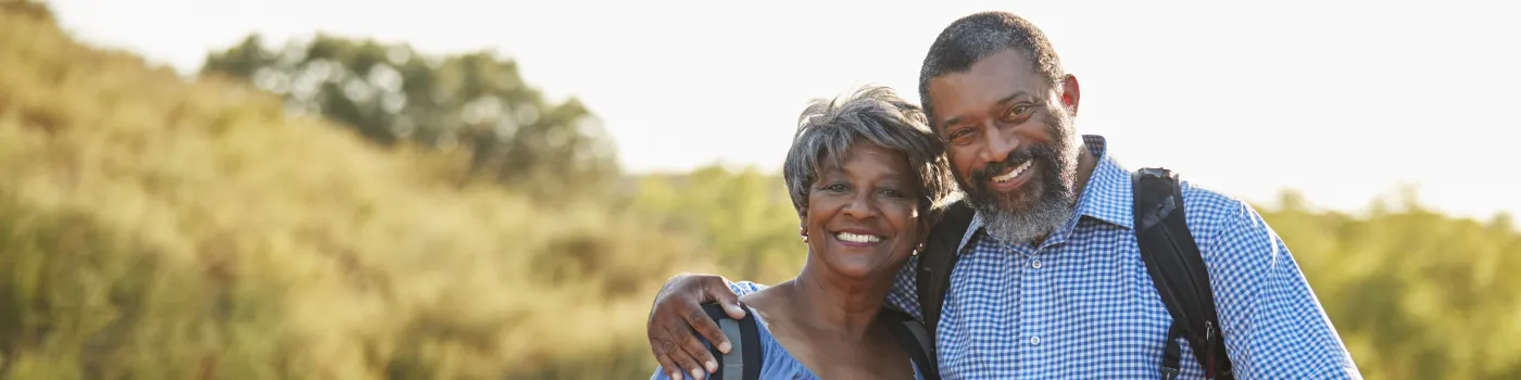 Black, elderly couple smiling with backpacks on as they hike. 