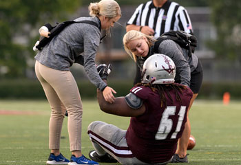 football player sitting on field be attending to by two sports medicine providers