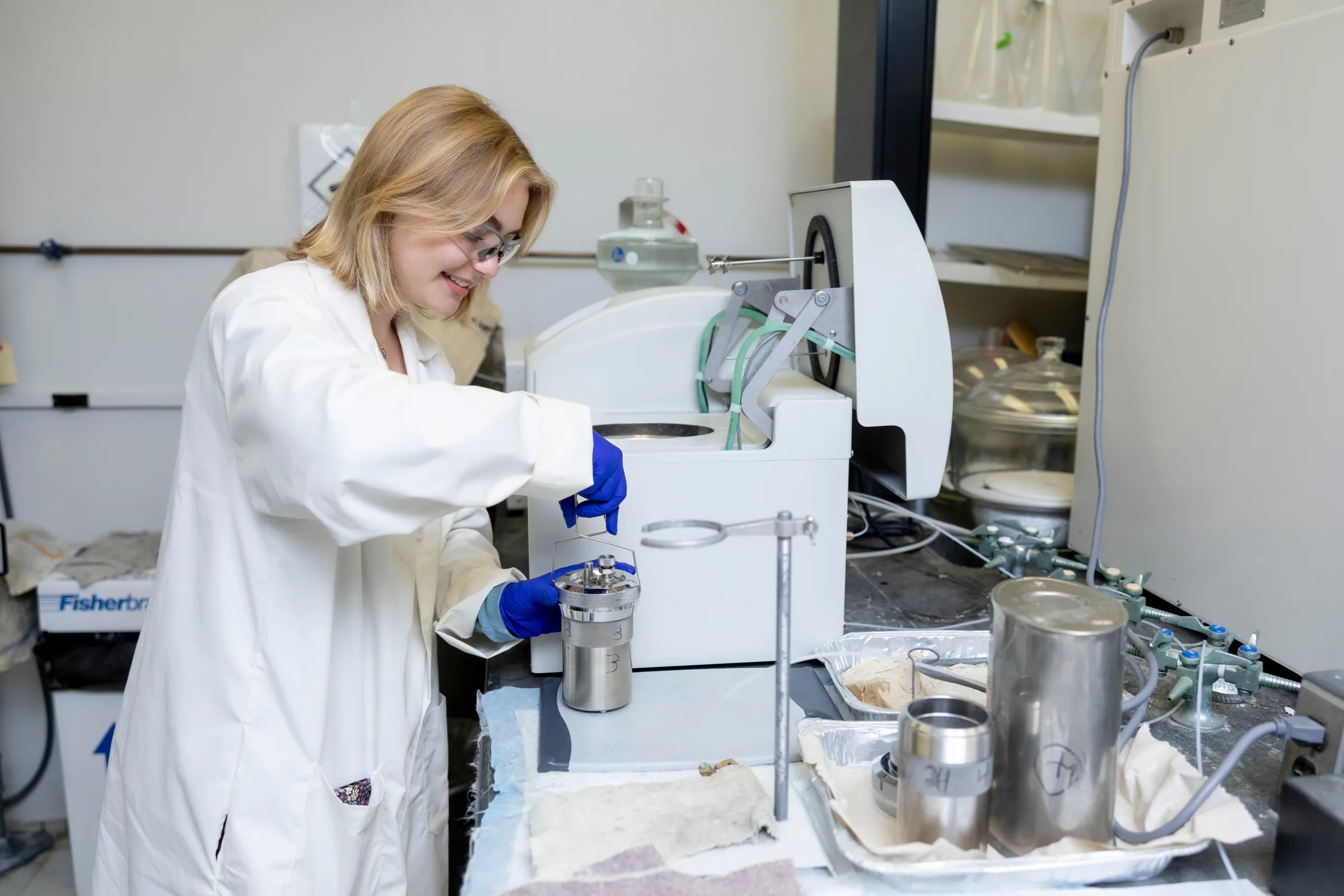 A woman in a white coat and goggles works at a lab station