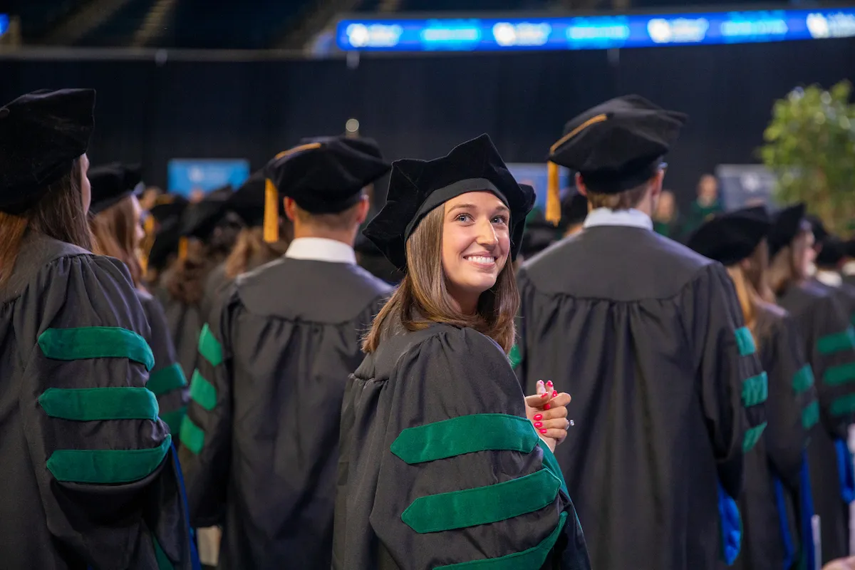 graduation ceremony for the glass of 2025. students standing in cap and gown with a female graduate looking back 