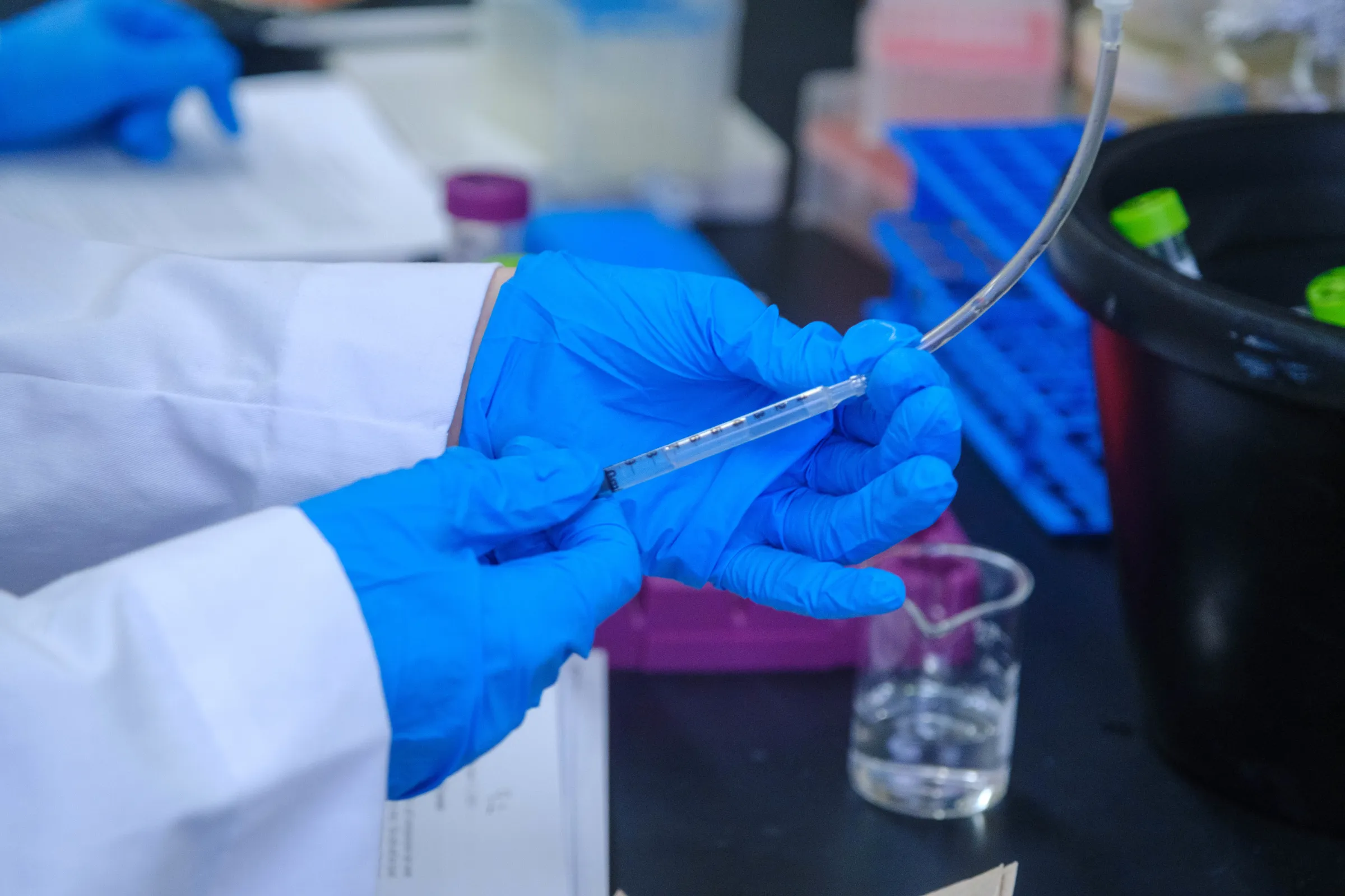 A researcher in a white coat and blue gloves handles a clear tube during a test