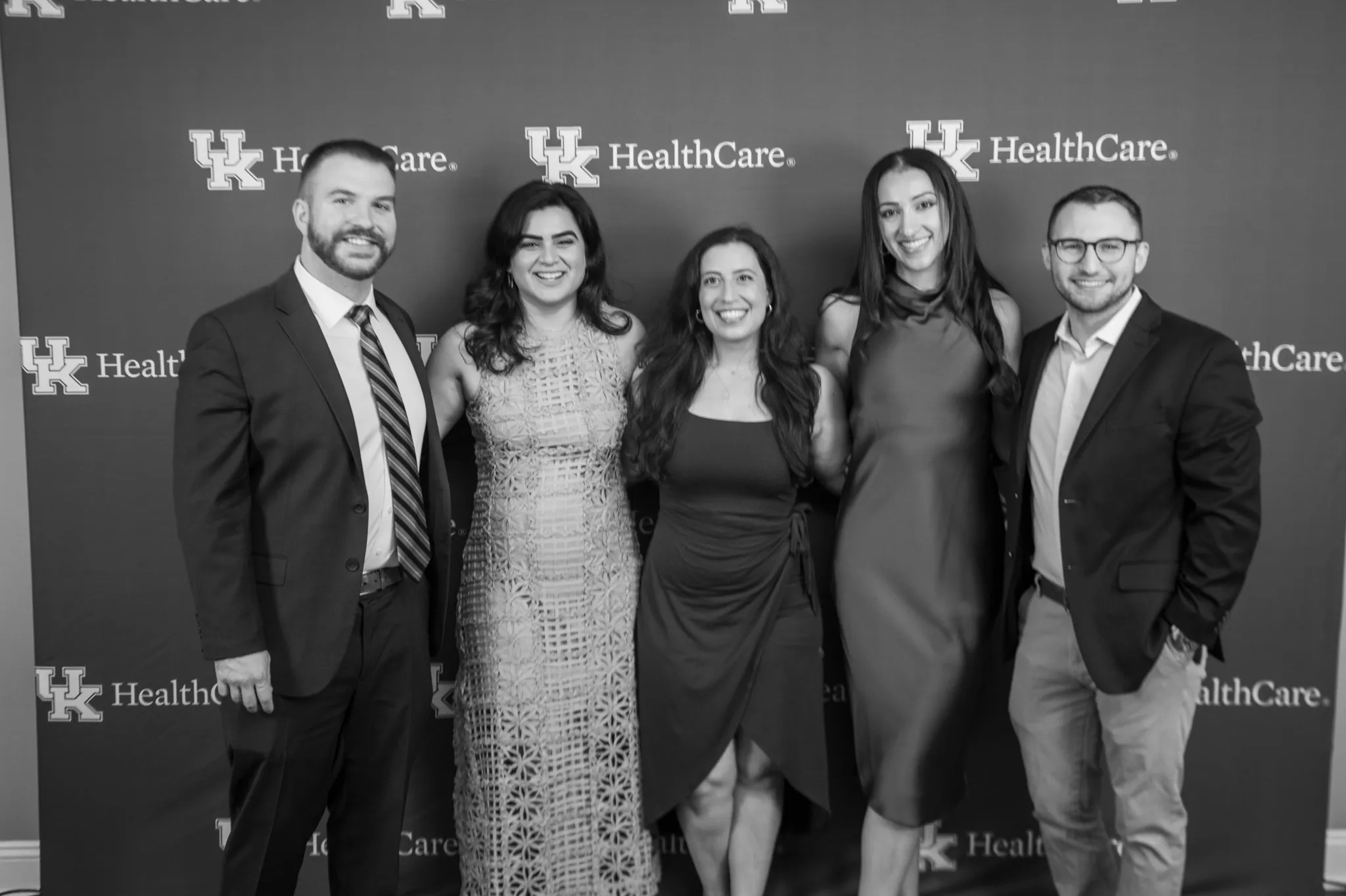A black and white photo of the program chiefs from last year, and the chiefs for next year gathered in front of a UK HealthCare wall.