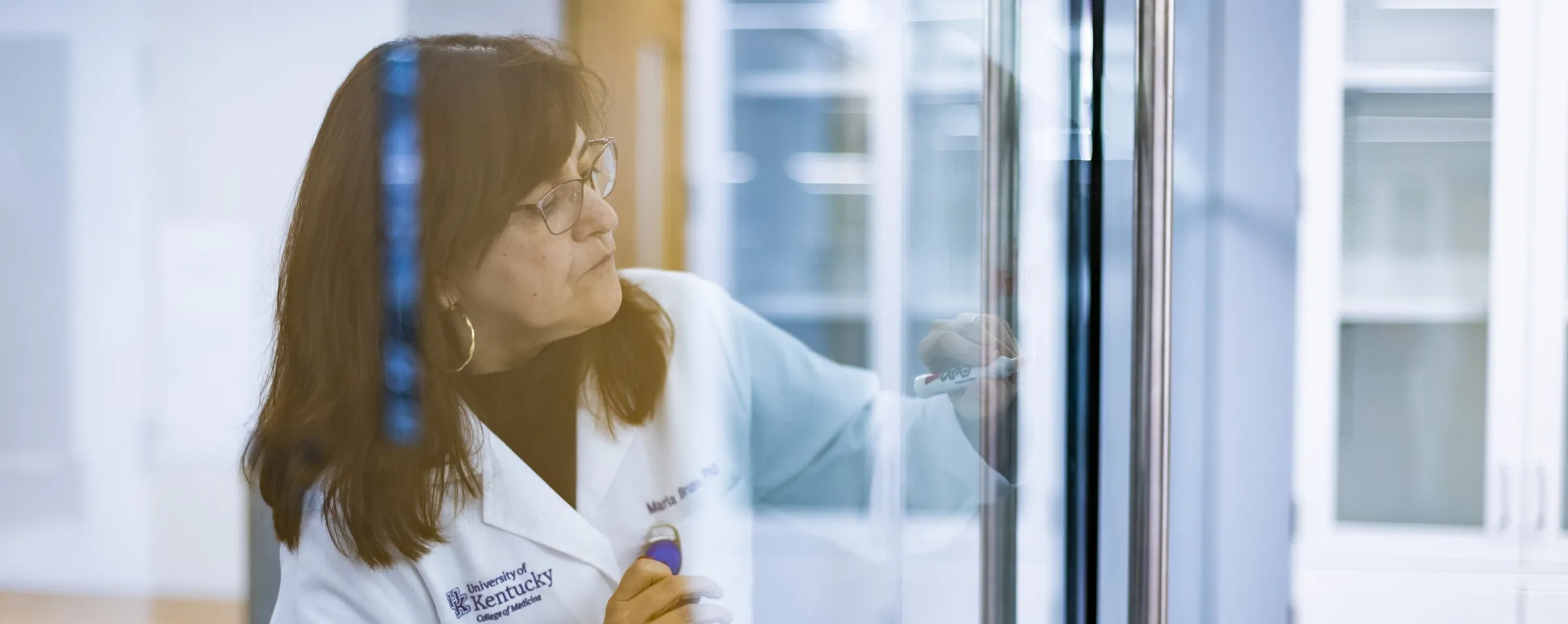 A woman in a lab coat writes on a see-through white board