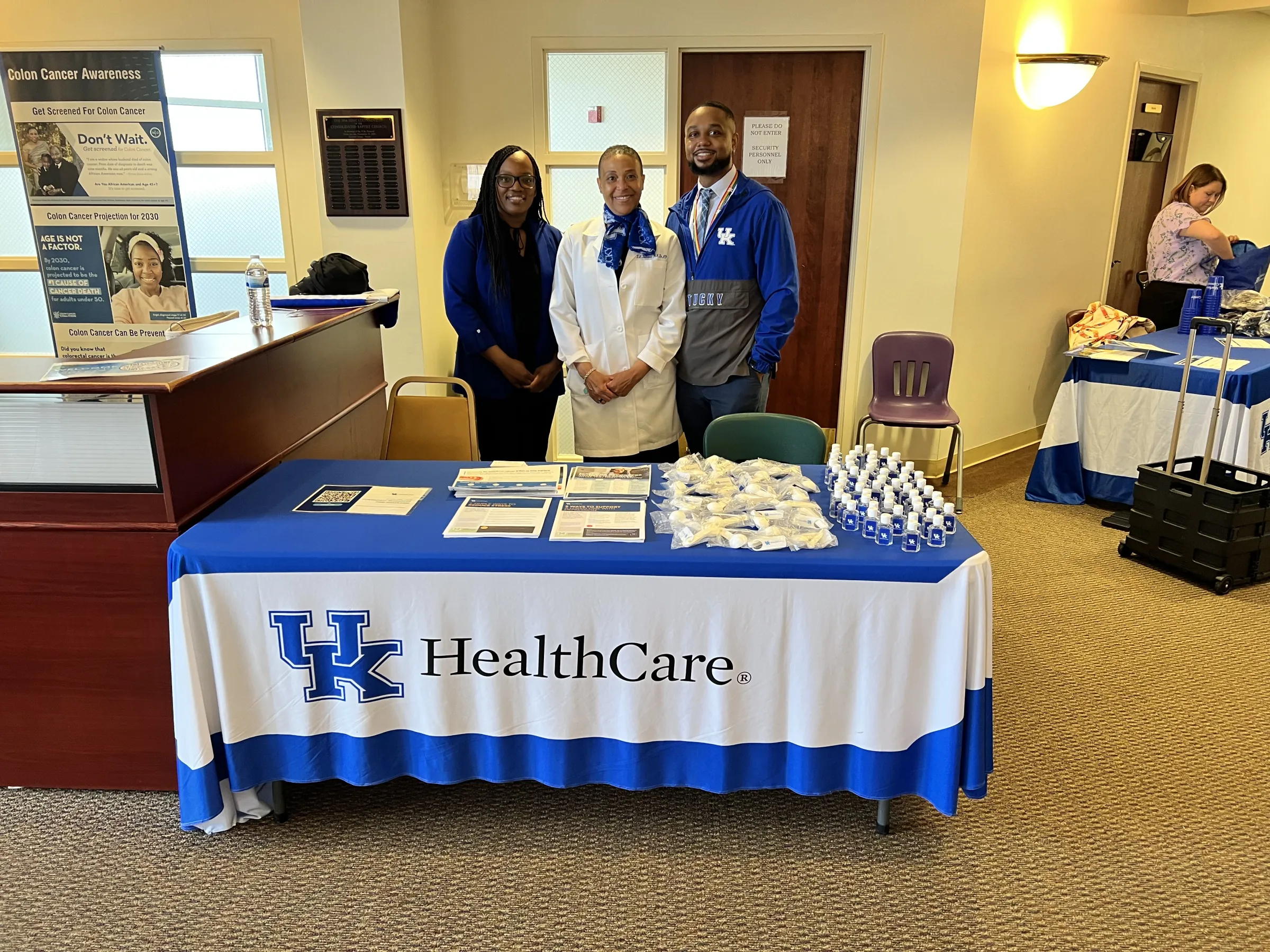 Three people stand smiling behind a swag table, labeled "UK HealthCare." 