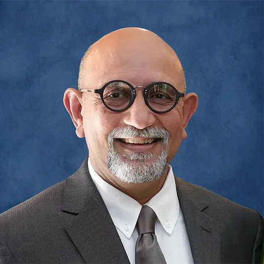 Dr. Ketan Buch, A man with round, dark-rimmed glasses, a grey coat, goatee, and a shaved head.