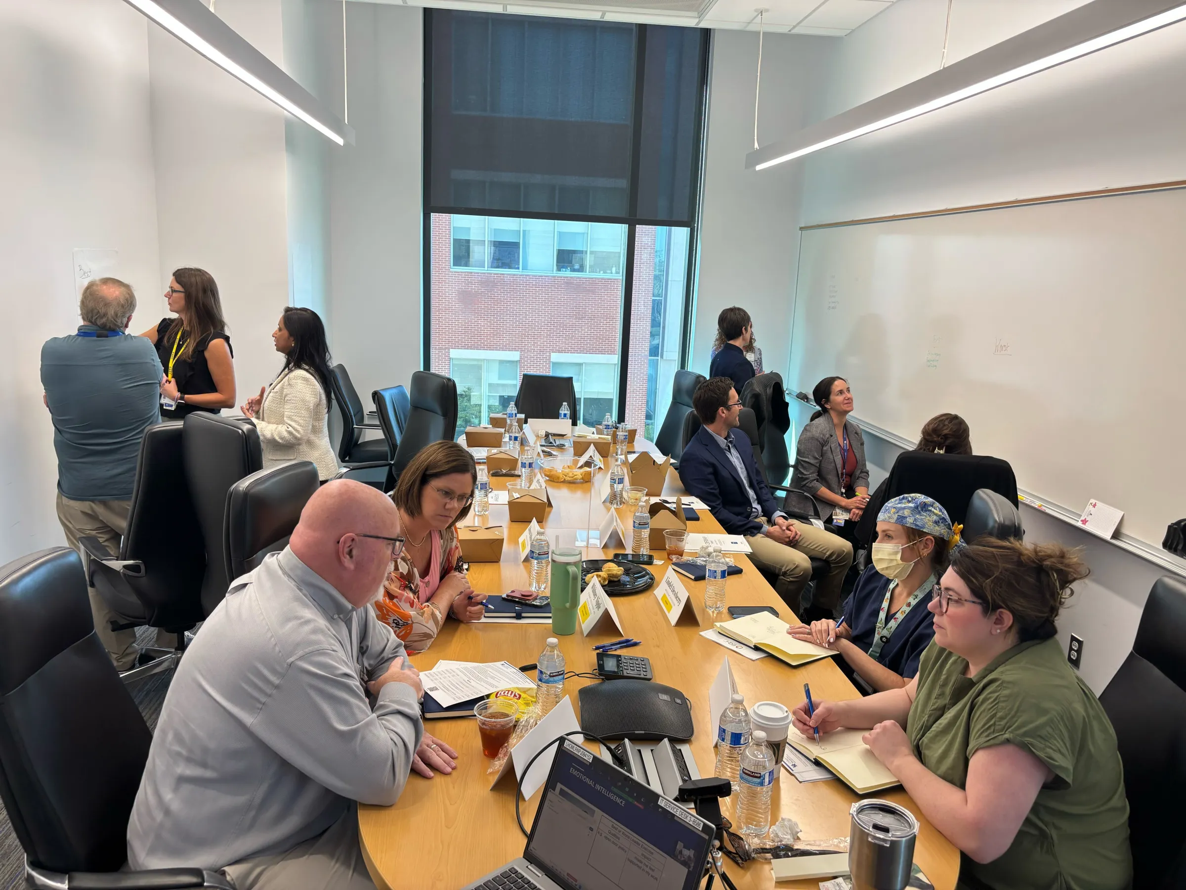 Standing and writing on white board with group of individuals talking with each other at a table.