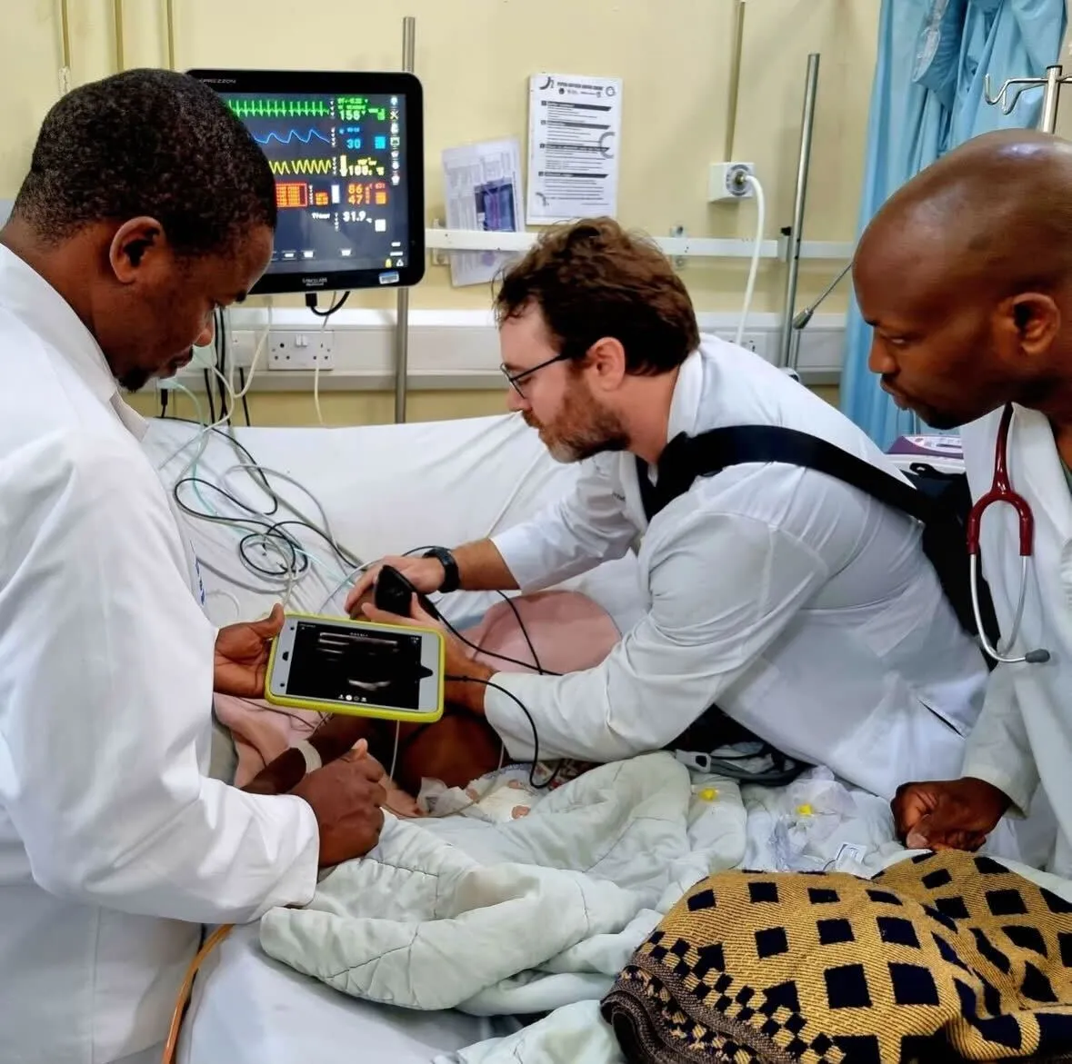 Physician Joel Hamm in ICU in Malawi (southeast Africa) assessing a pediatric patient while surrounded by other physicians and healthcare professionals who are assisting with ultrasound.
