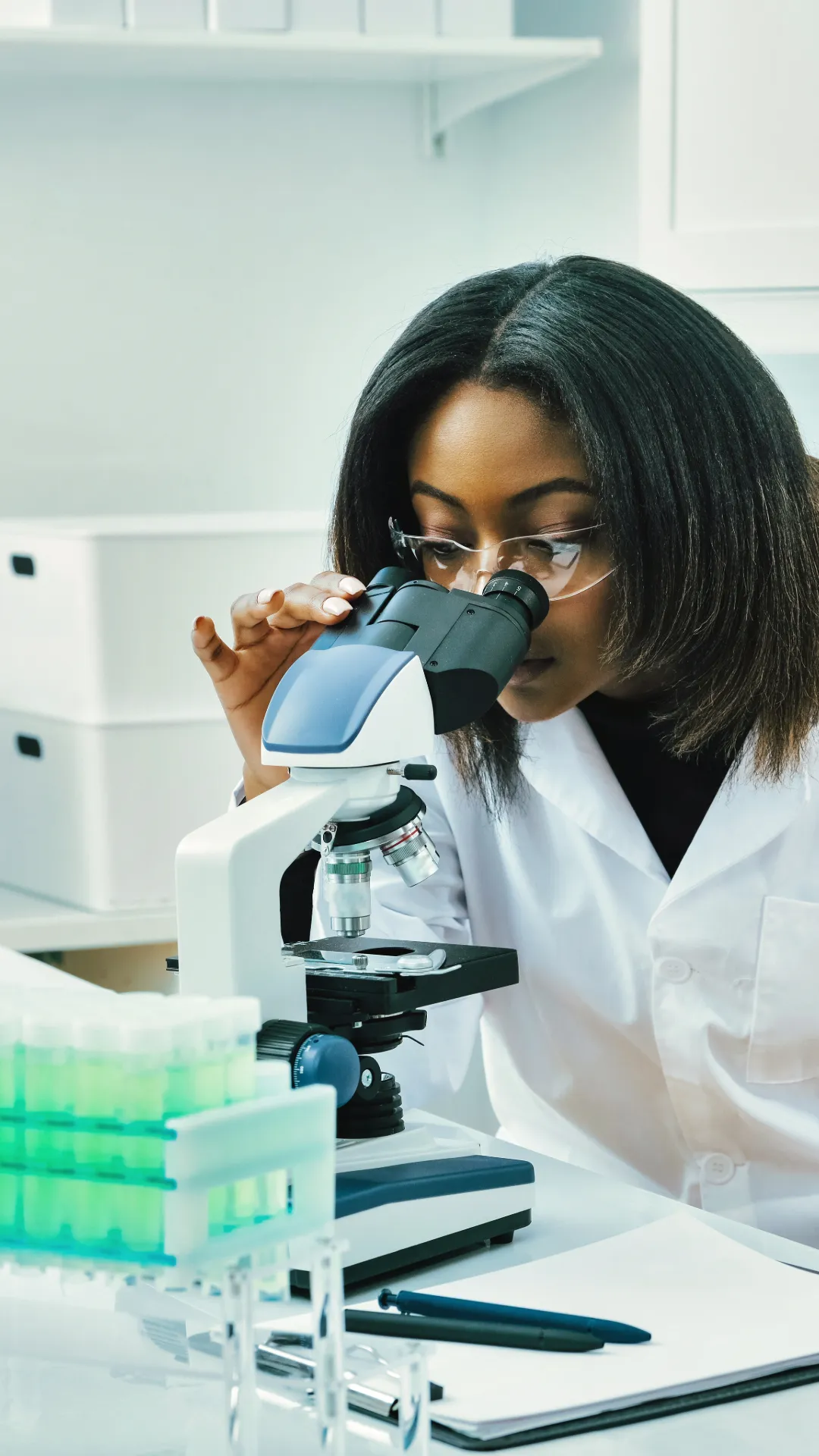 Woman in a lab looking into a microscope. On the desk, there are test tubes