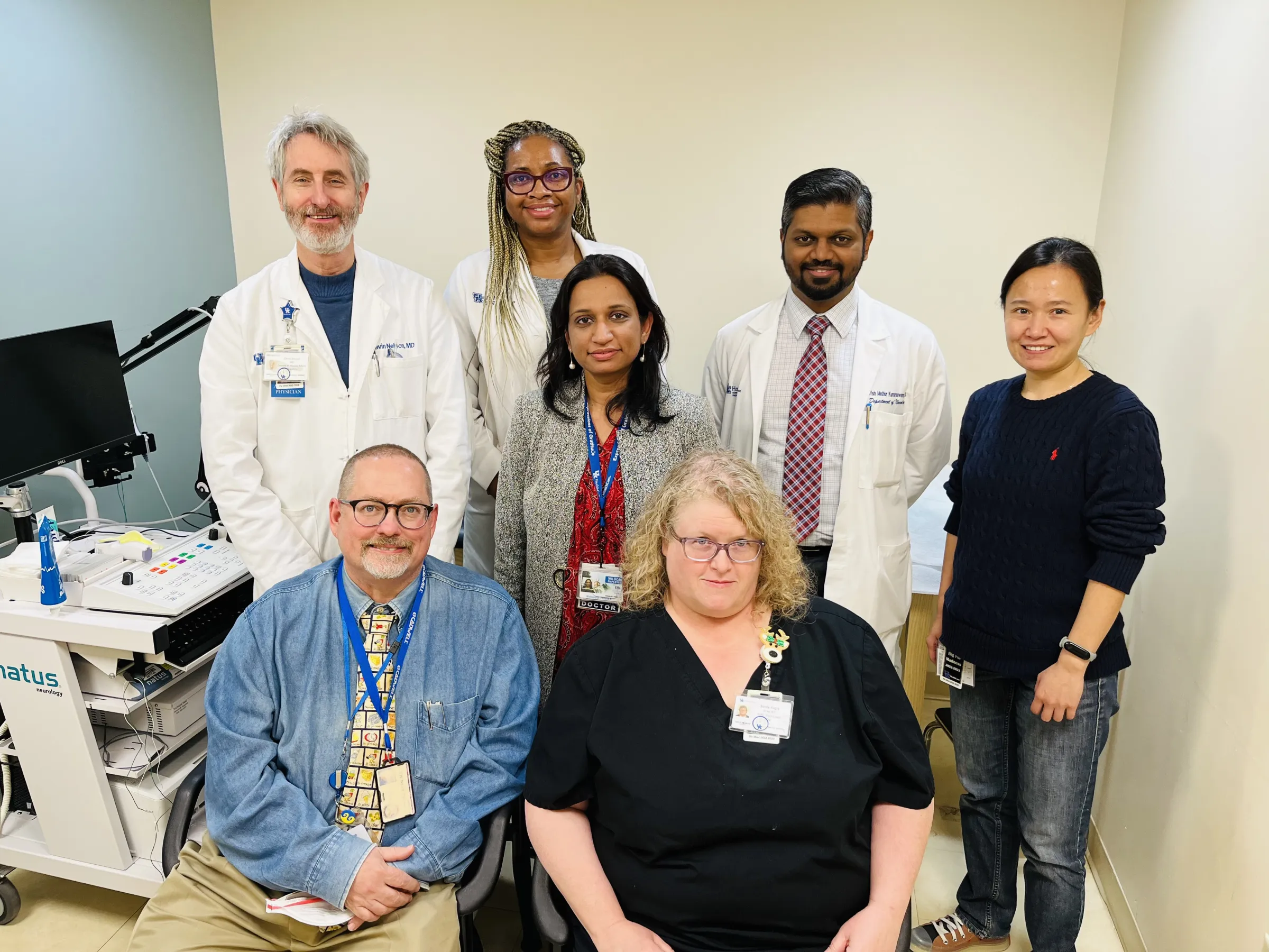 A diverse medical team of seven people, with four standing and three sitting, smiles at the camera in a clinical setting. They wear professional attire.