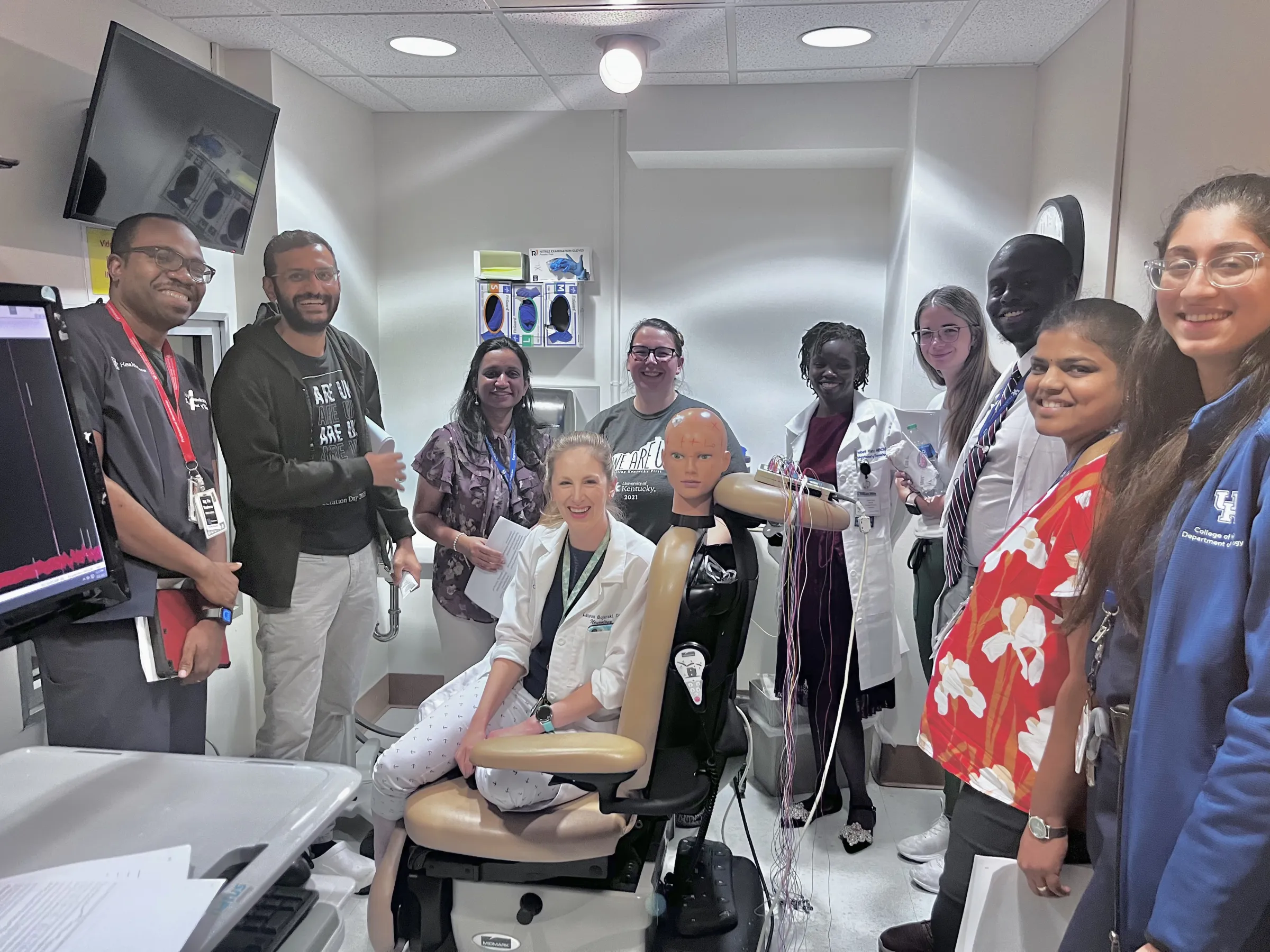 A group of diverse healthcare professionals pose smiling in a medical simulation room, gathered around a training mannequin in a dental chair, conveying teamwork and learning.