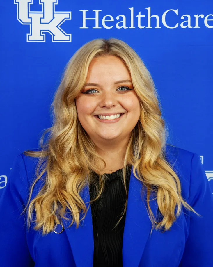 Christina Westfall, a blond woman wearing a UK blue blazer over a black shirt, stands in front of a UK Healthcare branded wall.