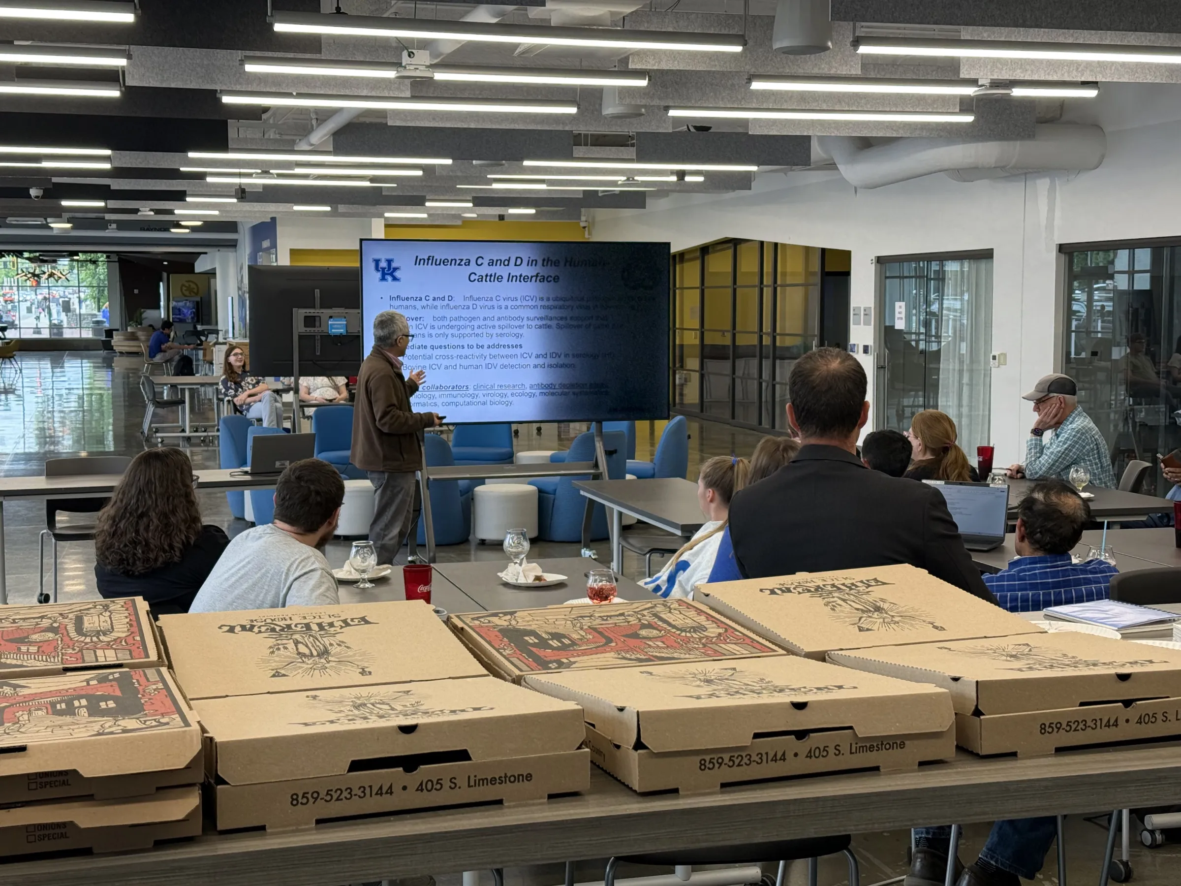 A group of people attend an informal research presentation in a modern lounge space. A presenter stands beside a large screen displaying a University of Kentucky slide titled “Influenza C and D in the Human-Cattle Interface.” Several pizza boxes, drinks, and laptops are visible on the tables in the foreground, suggesting a casual seminar or departmental gathering.
