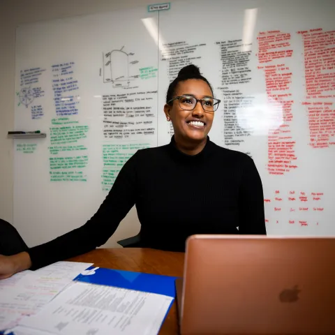 person sitting at desk with a whiteboard filled with writing behind her
