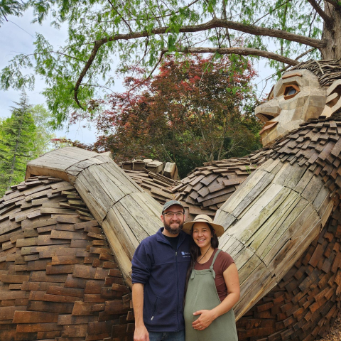 Shane Marsili standing with his pregnant wife at the Bernheim forest in front of a wooden sculpture of a giant.  