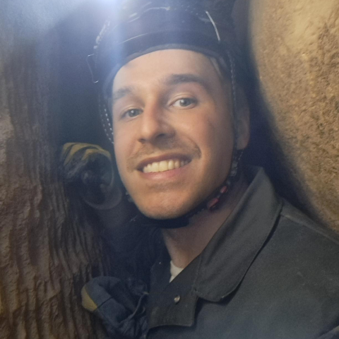 Clay Huffman smiling in-between two rocks in a cave with a miner light on his head. 