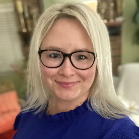 A blonde woman in a blue shirt and glasses smiles at the camera