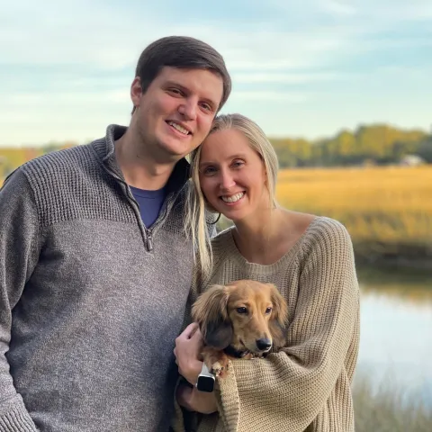 Cody Blanchard and his Wife smiling in front of a lake holding their long haired dachshund oscar