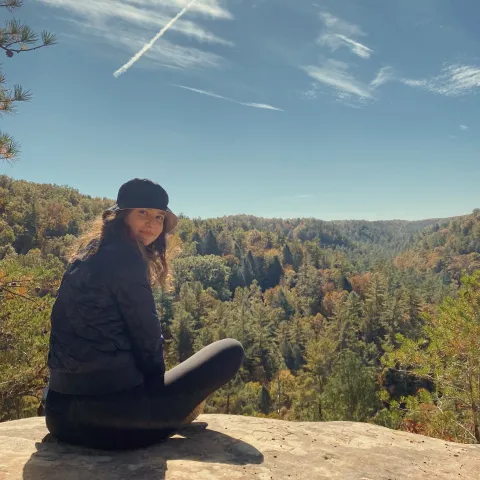 Diana Khair sitting at the top of a hiking lookout with the sky and tree landscape behind her. 