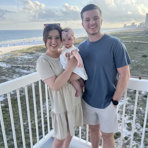 Anna Cox with her husband and baby daughter standing on a porch overlooking the beach.