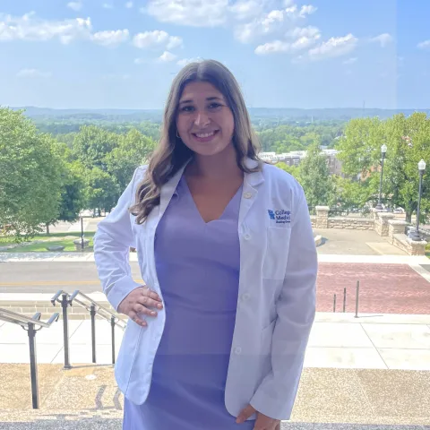 Student wearing white coat standing on stairs with city view in the background