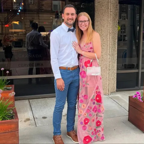 Thomas Calderaro and his significant other standing in front of a restaurant. She wears a floral dress and glasses, and he is in a white shirt and bow tie. Flower boxes flank them.
