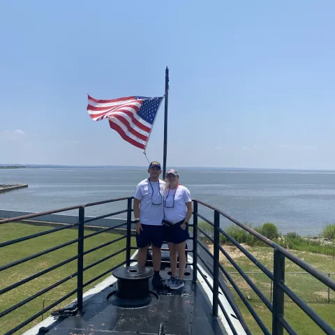 Zachary Combs and his wife stand on a ship deck under a sunny sky, with an American flag waving behind them. Calm water and grassy shoreline are visible in the background.