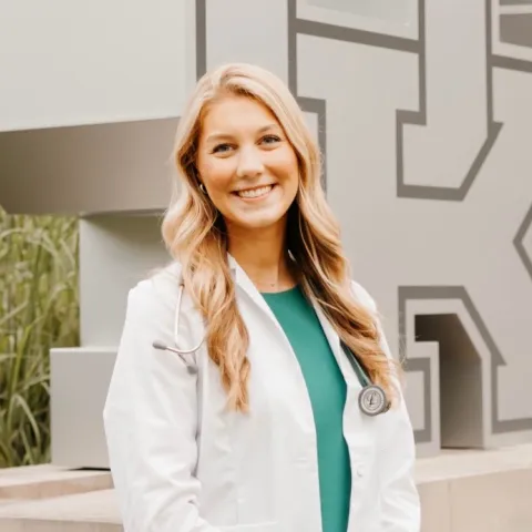 Student in white coat standing in front of UK statue.