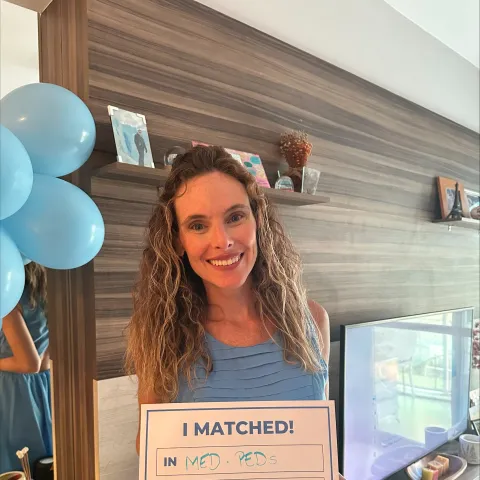 Luiza Goldemberg in a blue dress holds a sign reading "I matched in Med Peds at U Kentucky." Blue balloons and a cozy room setting are visible.