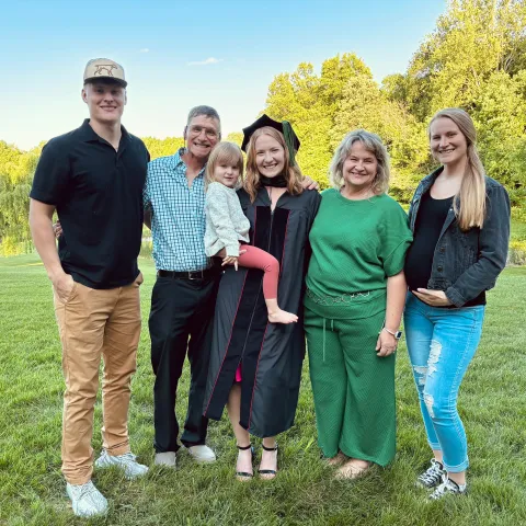 Anna Bowling and her happy family stands outdoors on grass with trees in the background. The young woman in a graduation gown and cap holds a child. Celebratory atmosphere.